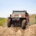 A muddy, red 4x4 off-roading vehicle driving over a mound of dirt with a green forest in the background.