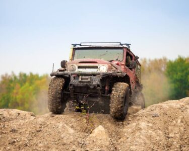 A muddy, red 4x4 off-roading vehicle driving over a mound of dirt with a green forest in the background.