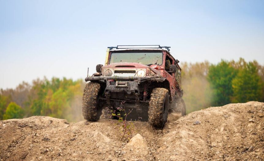 A muddy, red 4x4 off-roading vehicle driving over a mound of dirt with a green forest in the background.