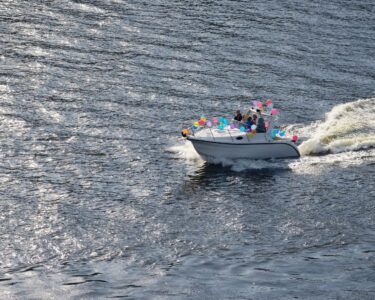 Aerial shot of a small boat out on the water with a wake behind it. Tied to the boat are several colorful balloons.