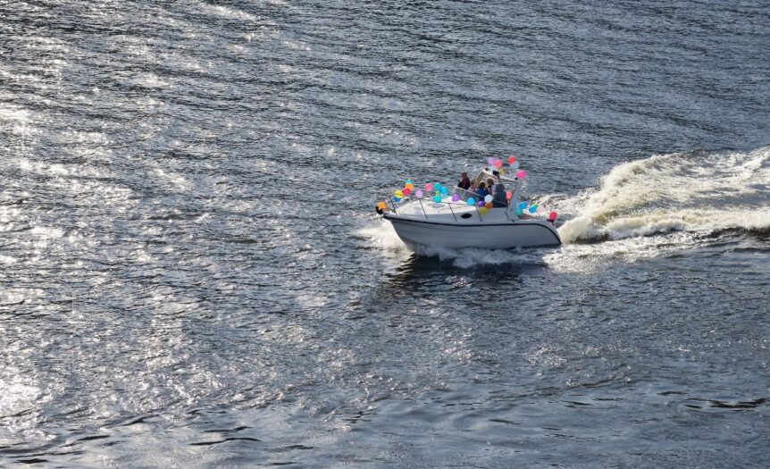 Aerial shot of a small boat out on the water with a wake behind it. Tied to the boat are several colorful balloons.