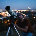 A man in a red shirt and glasses uses a telescope to look at the night sky with city lights blurred in the background.