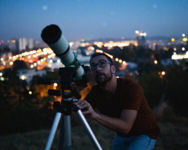 A man in a red shirt and glasses uses a telescope to look at the night sky with city lights blurred in the background.