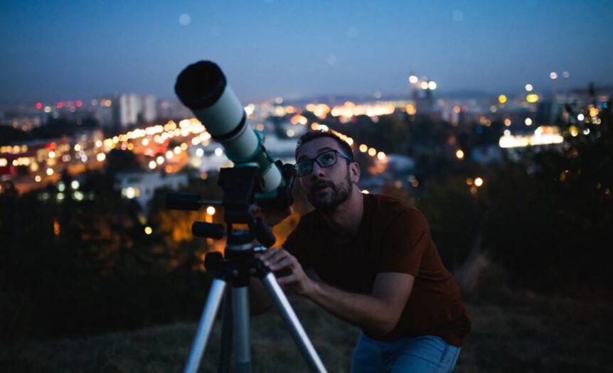 A man in a red shirt and glasses uses a telescope to look at the night sky with city lights blurred in the background.