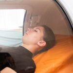 A young woman in a black top lying down on an orange bed in a hyperbaric oxygen chamber receiving oxygen therapy.