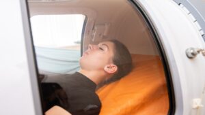 A young woman in a black top lying down on an orange bed in a hyperbaric oxygen chamber receiving oxygen therapy.