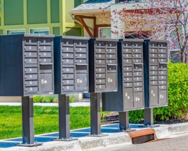 A row of cluster mailboxes stationed in front of a row of townhouses near a patch of grassy lawn and a tree.