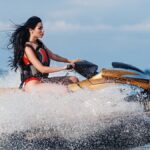 A dark-haired woman wearing a red life vest is riding a gold jet ski on the open water, making waves.