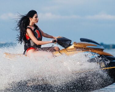 A dark-haired woman wearing a red life vest is riding a gold jet ski on the open water, making waves.