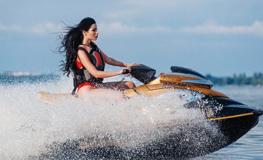 A dark-haired woman wearing a red life vest is riding a gold jet ski on the open water, making waves.