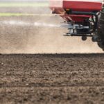 A close-up of a flat field of dirt. A tractor with a red fertilizer hopper on the back is spraying fertilizer across the dirt.