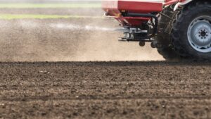 A close-up of a flat field of dirt. A tractor with a red fertilizer hopper on the back is spraying fertilizer across the dirt.