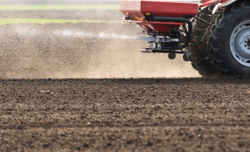 A close-up of a flat field of dirt. A tractor with a red fertilizer hopper on the back is spraying fertilizer across the dirt.