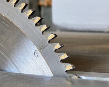 A close-up of a metal circular blade with large toothed edges. The blade is cutting a piece of metal.