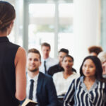 A close-up of a conference in session. There's a diverse group of attendees listening to the female keynote speaker.