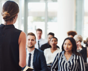 A close-up of a conference in session. There's a diverse group of attendees listening to the female keynote speaker.