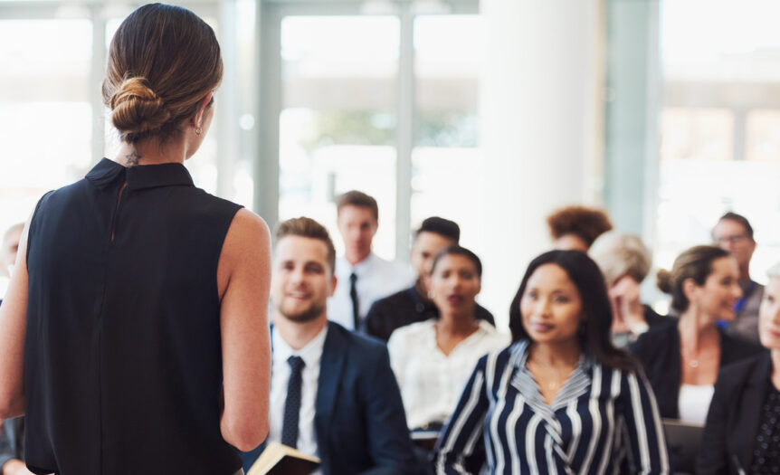 A close-up of a conference in session. There's a diverse group of attendees listening to the female keynote speaker.