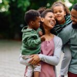 A young, loving family with two young children, one boy, and one girl, hanging out and smiling outside.