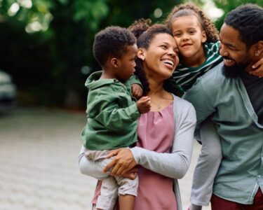 A young, loving family with two young children, one boy, and one girl, hanging out and smiling outside.