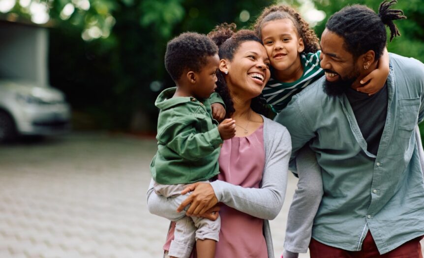 A young, loving family with two young children, one boy, and one girl, hanging out and smiling outside.