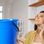 A woman stands in her home and holds a bucket while water drips into it from the ceiling. She has a cell phone on her ear.