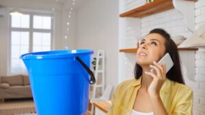 A woman stands in her home and holds a bucket while water drips into it from the ceiling. She has a cell phone on her ear.