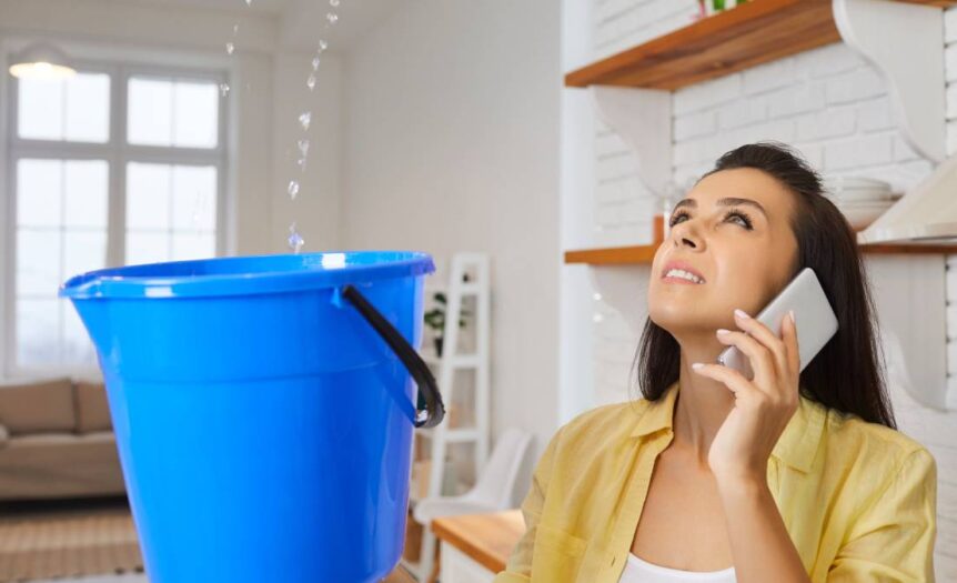 A woman stands in her home and holds a bucket while water drips into it from the ceiling. She has a cell phone on her ear.