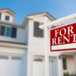 A red sign outside of a home labeled “For Rent.” It is a two-story house with a large garage and entryway.