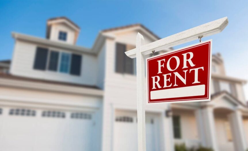 A red sign outside of a home labeled “For Rent.” It is a two-story house with a large garage and entryway.