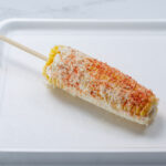 A white plate sits on a marble counter. Mexican street corn lies on the plate with a wooden skewer in the center.