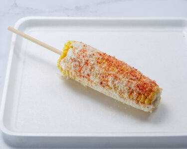 A white plate sits on a marble counter. Mexican street corn lies on the plate with a wooden skewer in the center.