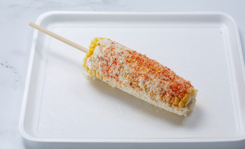 A white plate sits on a marble counter. Mexican street corn lies on the plate with a wooden skewer in the center.