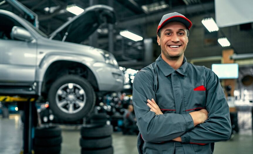 A smiling man wears a gray jacket and pants, crossing his arms, and standing in front of a car lift with a silver car on it.