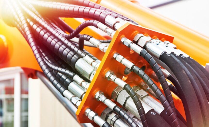 Several black hydraulic hoses attach to an orange piece of equipment with silver attachment. The sunshine hits the equipment.