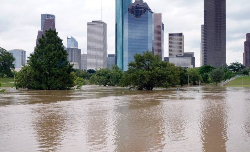 A flooded park lies in front of a city skyline. The water flooding the area is murky and brown.