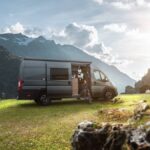 A dark grey-blue campervan that has been taken off-road to a grassy spot in the mountains with the sun shining in the background.