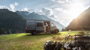 A dark grey-blue campervan that has been taken off-road to a grassy spot in the mountains with the sun shining in the background.