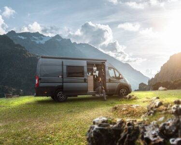 A dark grey-blue campervan that has been taken off-road to a grassy spot in the mountains with the sun shining in the background.