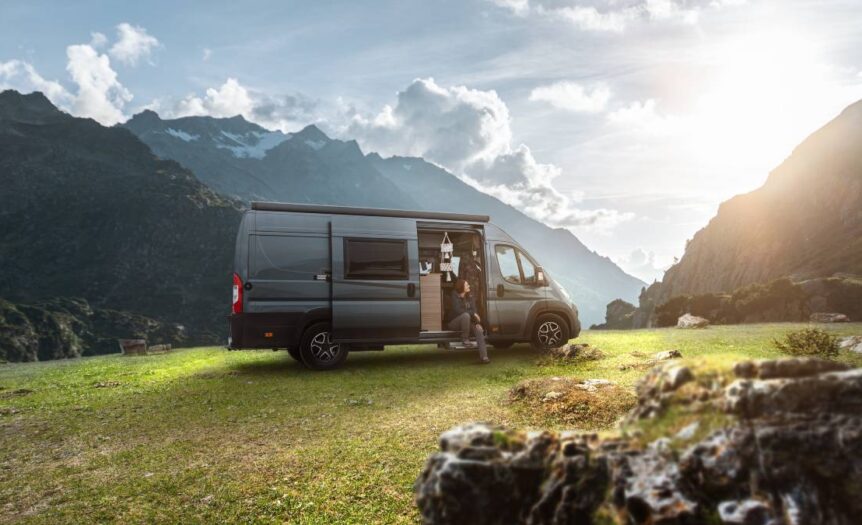 A dark grey-blue campervan that has been taken off-road to a grassy spot in the mountains with the sun shining in the background.