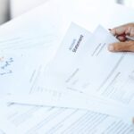 A personal banker sitting at the table with a pile of bank statements in front of them. Their black glasses are next to them.