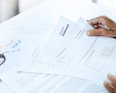 A personal banker sitting at the table with a pile of bank statements in front of them. Their black glasses are next to them.