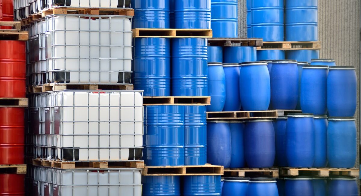 Stacks of chemical packaging containers, including metal barrels, plastic barrels, and plastic IBC tanks.