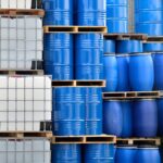 Stacks of chemical packaging containers, including metal barrels, plastic barrels, and plastic IBC tanks.