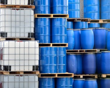 Stacks of chemical packaging containers, including metal barrels, plastic barrels, and plastic IBC tanks.