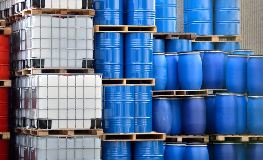 Stacks of chemical packaging containers, including metal barrels, plastic barrels, and plastic IBC tanks.