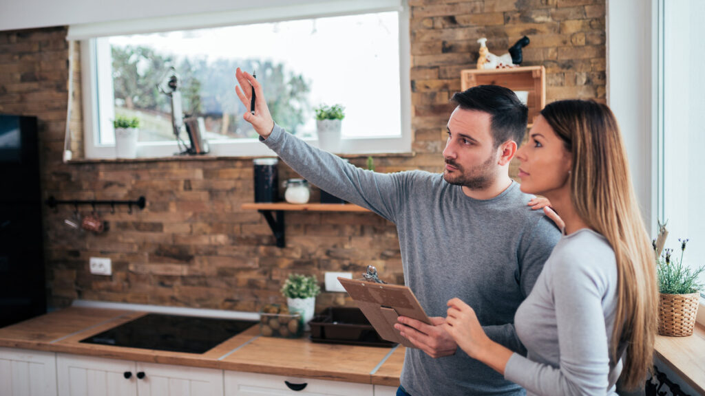 A young couple standing in their kitchen. The man is holding a clipboard and raising his hand while the woman looks on.