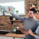 A young couple standing in their kitchen. The man is holding a clipboard and raising his hand while the woman looks on.