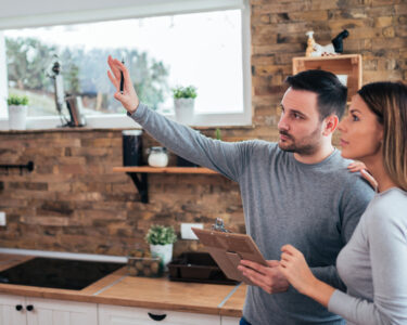 A young couple standing in their kitchen. The man is holding a clipboard and raising his hand while the woman looks on.