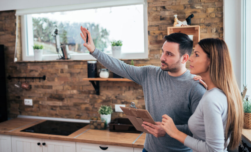 A young couple standing in their kitchen. The man is holding a clipboard and raising his hand while the woman looks on.