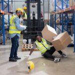 Two Black warehouse workers in safety vests. One worker fell over with two boxes falling on top of him.